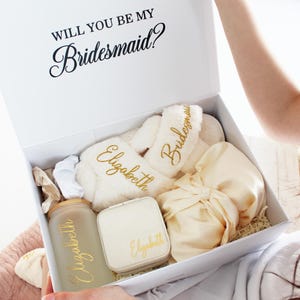 Może przedstawiać: Białe pudełko prezentowe z napisem "WILL YOU BE MY Bridesmaid?" zawiera różne przedmioty. Należą do nich puszyste białe kapcie z haftem "Elizabeth" i "Bridesmaid", szklany kubek z matowego szkła, małe pudełko na biżuterię i satynowa torebka.