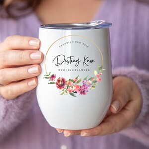 May include: White stainless steel wine tumbler with a gold circle and floral design. The text "Established 2018" and "Destiny Knox Wedding Planner" is printed on the tumbler.