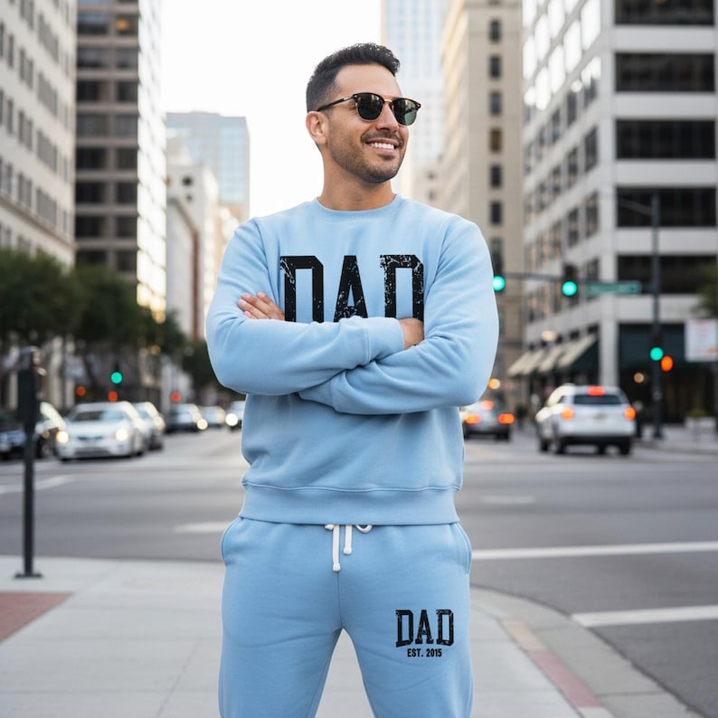 May include: A light blue sweatshirt and sweatpants set with the word "DAD" printed in black on the chest and leg. The sweatpants have "DAD EST. 2015" printed on the leg. The person is wearing sunglasses and standing on a city street.