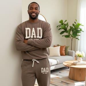 May include: A person wearing a brown sweatshirt and matching sweatpants. The sweatshirt and sweatpants have the word "DAD" printed on them in a distressed white font. The sweatshirt also has "EST. 2022" printed below the word "DAD".