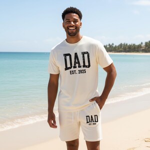 May include: Cream-colored t-shirt and shorts set with the text "DAD EST. 2025" in a distressed black font. The matching shorts have a drawstring waist. The person is standing on a sandy beach with the ocean in the background.