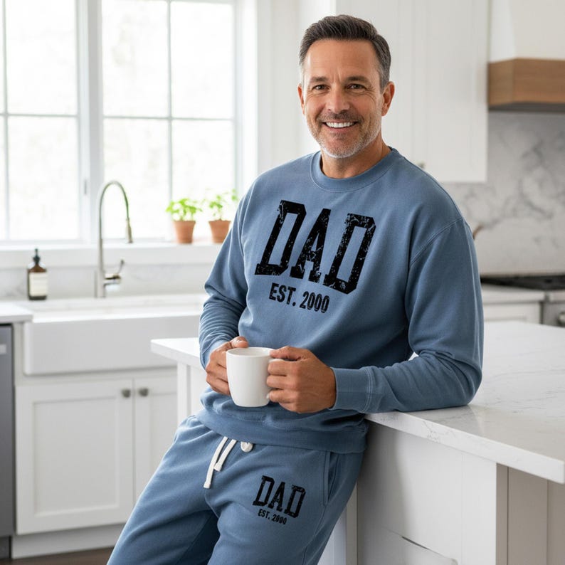May include: A light blue sweatshirt and sweatpants set with the word "DAD" and "EST. 2000" printed in black. The man is holding a white mug. The set is casual and comfortable.