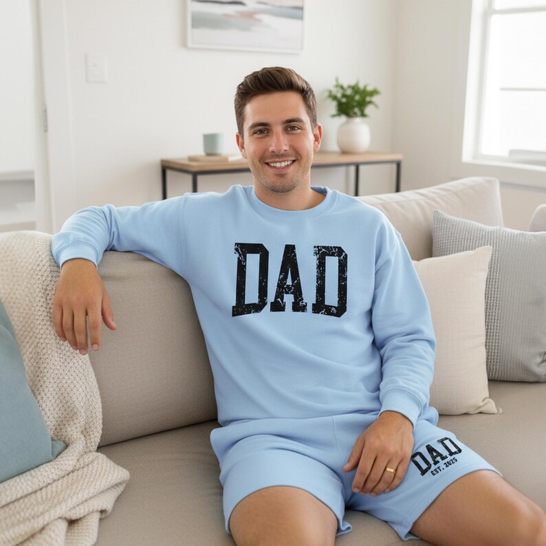 May include: A light blue sweatshirt and shorts set. The sweatshirt features the word "DAD" in a distressed black font. The shorts also have "DAD" printed on them, along with "EST. 2023". The set is made of a soft material.