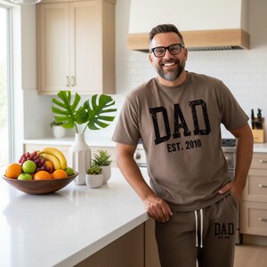 May include: A man wearing a brown t-shirt and matching sweatpants. The t-shirt has the word "DAD" printed in black, with "EST. 2010" below it. The man is smiling and wearing glasses. A wooden bowl of fruit is on the counter.