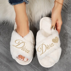 May include: White fluffy slippers with a crisscross design. The word "Diva" is written in gold glitter on each slipper. One slipper has a gold glitter heart. The slippers are on a gray surface.