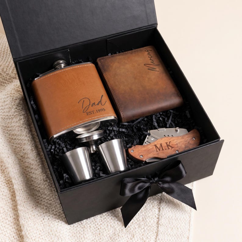 K&ouml;nnte beinhalten: Schwarze Geschenkbox mit einer braunen Lederflasche, einem Portemonnaie, einem Taschenmesser und zwei Schnapsgl&auml;sern. Die Flasche ist mit "Dad EST.1995" graviert. Das Portemonnaie ist mit einem Namen graviert. Das Taschenmesser hat einen Holzgriff und die Initialen "M.K."