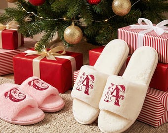 Può includere: Un paio di soffici pantofole natalizie bianche con un design a lettera "A" rossa e omino di pan di zenzero. Sono visibili anche un paio di pantofole rosa. Le pantofole sono vicino a regali confezionati e un albero di Natale decorato.