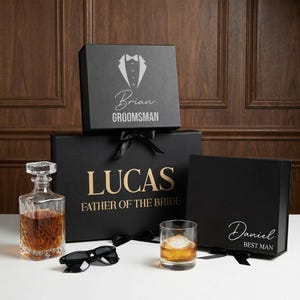 May include: A collection of black gift boxes with white and gold lettering, and a tuxedo graphic. One box reads "Brian Groomsman", another "Lucas Father of the Bride", and a third "Daniel Best Man". Also, a decanter, a glass of whiskey, and sunglasses.