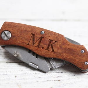 K&ouml;nnte beinhalten: Ein Holz-Utility-Messer mit einer Edelstahlklinge. Das Messer hat einen dunkelbraunen Holzgriff mit den eingravierten Initialen "M.K.". Die Klinge ist teilweise ausgefahren und zeigt die scharfe Kante und einen transparenten Kunststoff-Klingenhalter.