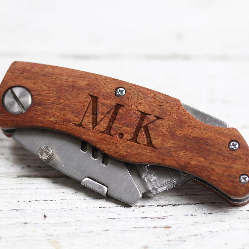 K&ouml;nnte beinhalten: Ein Holz-Utility-Messer mit einer Edelstahlklinge. Das Messer hat einen dunkelbraunen Holzgriff mit den eingravierten Initialen "M.K.". Die Klinge ist teilweise ausgefahren und zeigt die scharfe Kante und einen transparenten Kunststoff-Klingenhalter.
