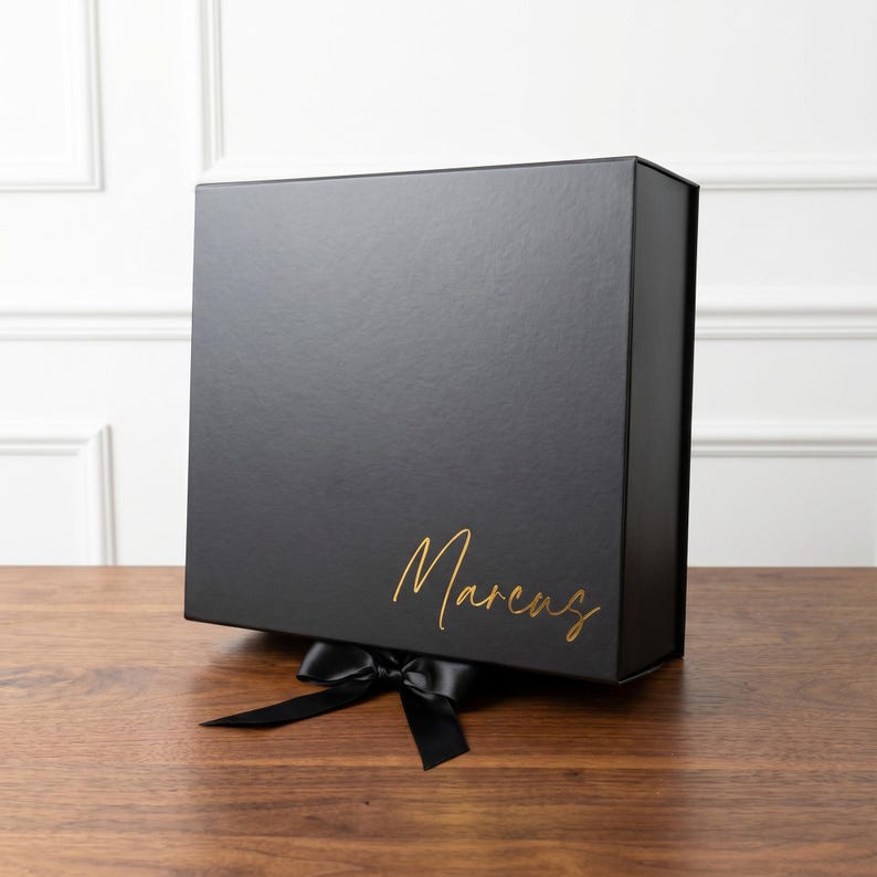 A black gift box with a matching black satin ribbon and bow. The box lid features the name "Marcus" in gold script. The box is sitting on a light wood surface.