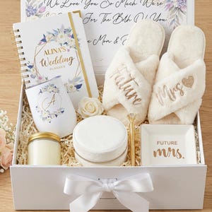 May include: A white gift box filled with wedding-themed items. Includes a planner, tumbler, candle, jewelry box, pen, slippers with "Future Mrs" embroidery, and a small dish. Floral and gold accents are present throughout the items.