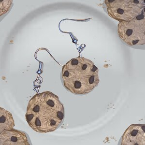 Könnte beinhalten: Ein Paar silberne Ohrringe mit Haken und Anhängern in Form von Schokoladen-Chip-Cookies. Die Cookies sind braun mit dunkelbraunen Schokoladenstückchen.