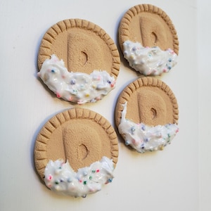 May include: Four round, tan-colored cookies with a textured edge and a central indentation. Each cookie is partially dipped in white frosting with colorful sprinkles, creating a sweet treat appearance. The cookies are arranged on a white surface.