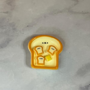 May include: A small, yellow and white bread-shaped charm with three pieces of toast and a square of butter. The charm has a glossy finish and is likely made of resin or a similar material. The charm is a food-themed accessory.