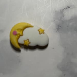 May include: A charming embellishment depicting a yellow crescent moon with a pink cheek and a white cloud with three yellow stars. The charm is set against a marbled background.