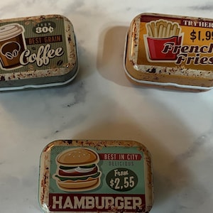 May include: Three rectangular metal tins with vintage-style food graphics. One tin features a coffee cup and the word "Coffee". Another tin displays a container of French fries and the words "French Fries". The third tin shows a hamburger and the word "Hamburger".