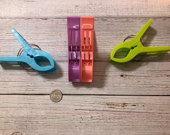May include: Three plastic clothespins in different colors: light blue, purple, and green. Each clothespin has a metal spring.