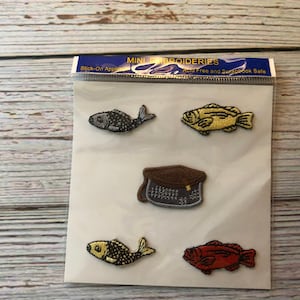 May include: Five embroidered iron-on patches, including a gray fish, a yellow fish, a brown and gray hat, a yellow and black fish, and a red fish. The patches are on a white background with the text "MINI EMBROIDERIES Acid Free and Scrapbook Safe" at the top.