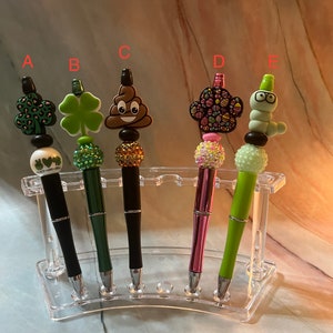 May include: Five pens with different colorful and decorative tops in a clear plastic pen holder. The tops include a green shamrock, a brown poop emoji, a pink and green floral design, a green worm with glasses, and a green shamrock with black spots.