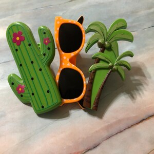 May include: A set of three colorful plastic decorations featuring a green cactus with pink flowers, a pair of orange sunglasses with white polka dots, and a green palm tree with brown trunk.