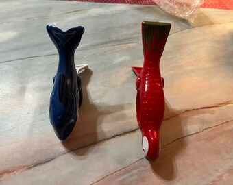 May include: Two fish-shaped clothespins. One is navy blue with a white tail design, and the other is red with green accents on the tail. Both have a white clip.