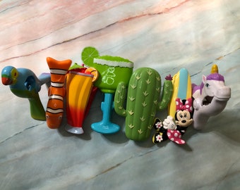 May include: A set of colorful plastic toys featuring a parrot, a clownfish, a margarita glass, a cactus, a surfboard, a Minnie Mouse figure, and a unicorn. The toys are all in a playful and whimsical style.