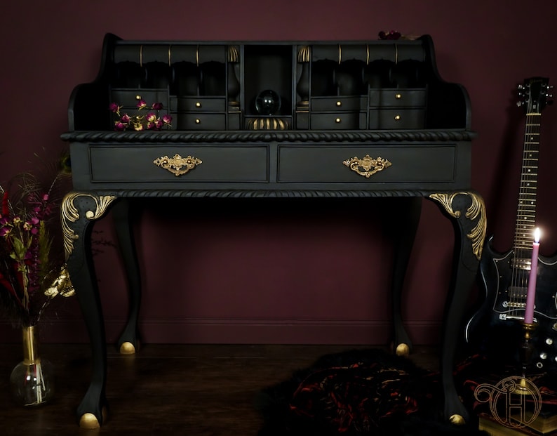 May include: A black writing desk with gold accents and intricate details. The desk has two drawers and a top section with multiple compartments. The desk is on a dark wood floor.