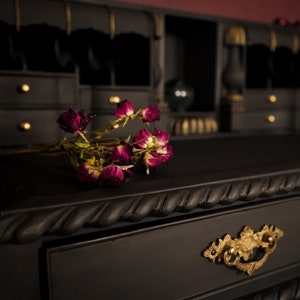 May include: A black wooden desk with gold accents and a drawer with an ornate gold handle. Dried roses are placed on the desk surface.