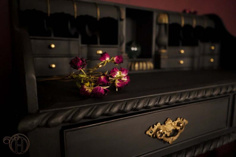 May include: A black wooden desk with gold accents and a drawer with an ornate gold handle. Dried roses are placed on the desk surface.