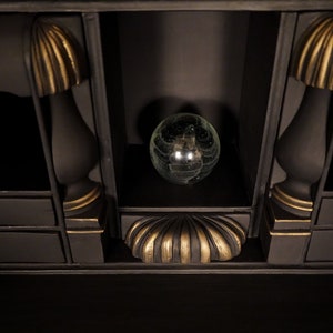 May include: A black wooden cabinet with gold accents and a glass orb on the top shelf. The cabinet has two drawers with gold knobs.