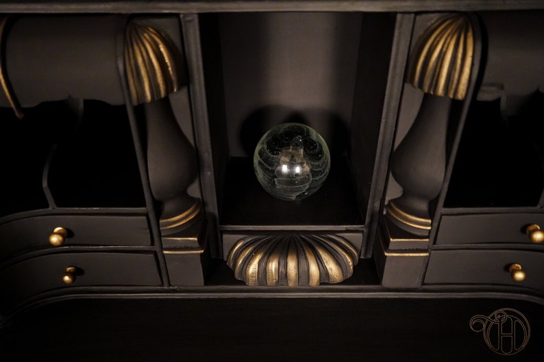 May include: A black wooden cabinet with gold accents and a glass orb on the top shelf. The cabinet has two drawers with gold knobs.