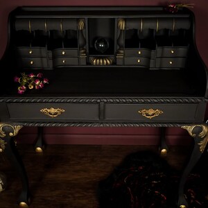 May include: A black writing desk with gold accents and two drawers. The desk has a raised top section with a small shelf and multiple compartments. The desk is sitting on a wooden floor with a guitar and a candle in the foreground.