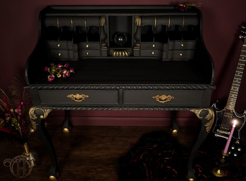 May include: A black writing desk with gold accents and two drawers. The desk has a raised top section with a small shelf and multiple compartments. The desk is sitting on a wooden floor with a guitar and a candle in the foreground.