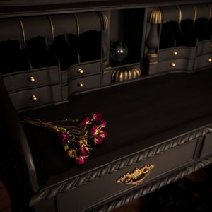 May include: A black writing desk with gold accents and a drawer with a rope-like design. The desk has a top surface with a small bouquet of dried roses.
