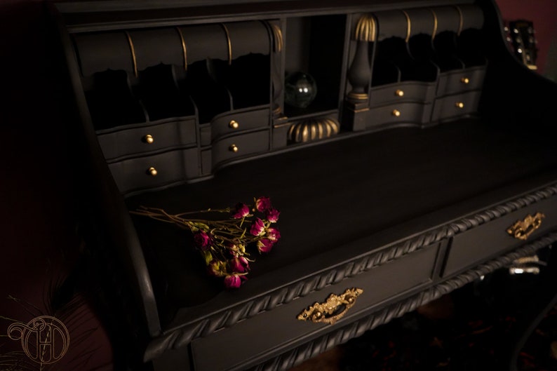 May include: A black writing desk with gold accents and a drawer with a rope-like design. The desk has a top surface with a small bouquet of dried roses.