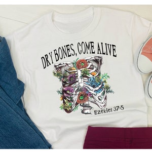 May include: White t-shirt with a black and white illustration of a ribcage with flowers growing inside. The text "Dry Bones, Come Alive" is above the illustration and "Ezekiel 37:5" is below.