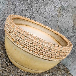 Pode incluir: Uma tigela redonda de cerâmica bege com interior liso e fosco e um padrão texturizado em forma de grade ao redor da borda superior. A superfície externa apresenta um acabamento sutil em tom castanho claro, com um design em relevo.