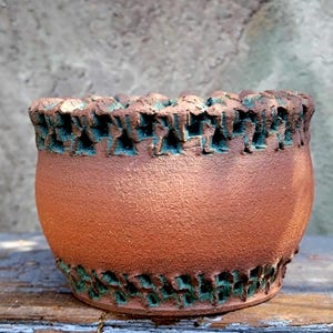 May include: A handcrafted, textured ceramic bowl in shades of brown and teal. The bowl features a rough, earthy texture with a decorative teal pattern around the rim and base. The bowl is suitable for holding small plants or decorative items.