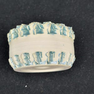 May include: A handmade, cylindrical ceramic container with a light cream base and a textured, turquoise blue rim and base. The container has a smooth, unadorned central band. The turquoise blue color is repeated in the textured pattern around the rim and base.