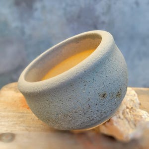 May include: A small, light blue ceramic bowl with a textured surface. The bowl is filled with a yellow liquid. The bowl is tilted slightly, revealing its contents. The bowl is sitting on a wooden surface.