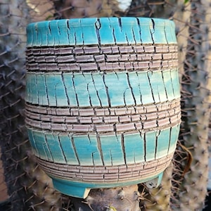 May include: A turquoise and brown ceramic planter with a textured, cracked glaze. The cylindrical pot features horizontal bands of brown and turquoise, with a small base and a slightly flared rim. The planter is designed for indoor or outdoor use.