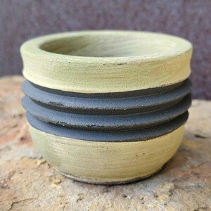 May include: A small, handmade ceramic pot with a textured, light green exterior and a dark gray, ridged band around the middle. The pot has a rounded base and a wide, open top, suitable for small plants or decorative use.