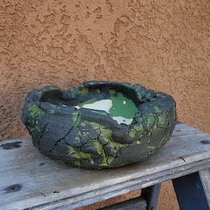 May include: A handcrafted, textured ceramic bowl with a dark, weathered exterior and green accents. The interior features a white and green painted surface. The bowl sits on a wooden surface, showcasing its unique, organic design.