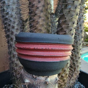 Może przedstawiać: Czarny i czerwony ceramiczny pojemnik na rośliny z żebrowanym wzorem. Pojemnik stoi na tle dużego kaktusa z ostrymi kolcami. Doniczka jest okrągła i ma teksturowaną powierzchnię.