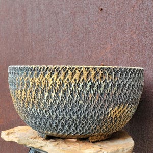 May include: A textured, ceramic bowl with a dark gray and yellow color scheme. The bowl has a rough, pitted surface and sits on a natural stone. The background is a rusty brown color.
