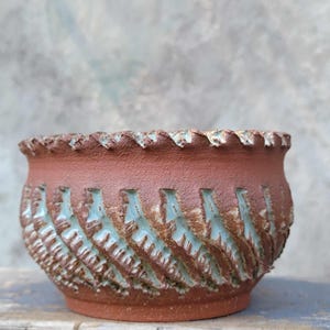 May include: A handcrafted, terracotta-colored ceramic planter with a textured surface. The rim has a scalloped edge, and the body features a repeating pattern of carved, angled lines with a turquoise-colored interior. The planter is suitable for indoor or outdoor use.