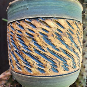 Puede incluir: Una maceta de cerámica hecha a mano con un diseño tallado y texturizado. La maceta presenta una banda de arcilla marrón dorada texturizada con detalles en azul, enmarcada por bandas de esmalte verde-azulado. La maceta es cilíndrica.