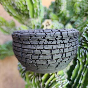 May include: A small, dark gray ceramic bowl with a textured, cracked surface. The bowl is round and sits in front of a blurred background of green plants. The bowl's exterior has a rough, weathered appearance.