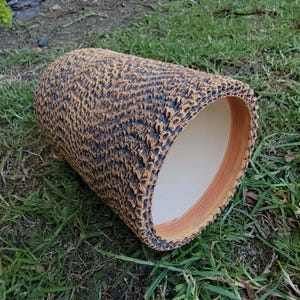May include: A cylindrical ceramic planter with a textured, bark-like exterior in shades of brown and black. The interior is a light beige color, and the rim has a reddish-brown accent. The planter is resting on green grass.
